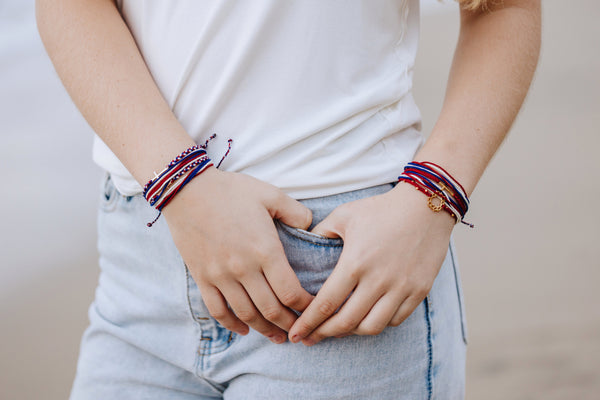 Christian model wearing rose gold cross bracelet hand braided in red, white, blue cotton cord, engraved "WE TRUST IN GOD" with Rizen Jewelry and Made 4 Ministries round disc tag. and multiple other red, white and blue cross bracelets. 