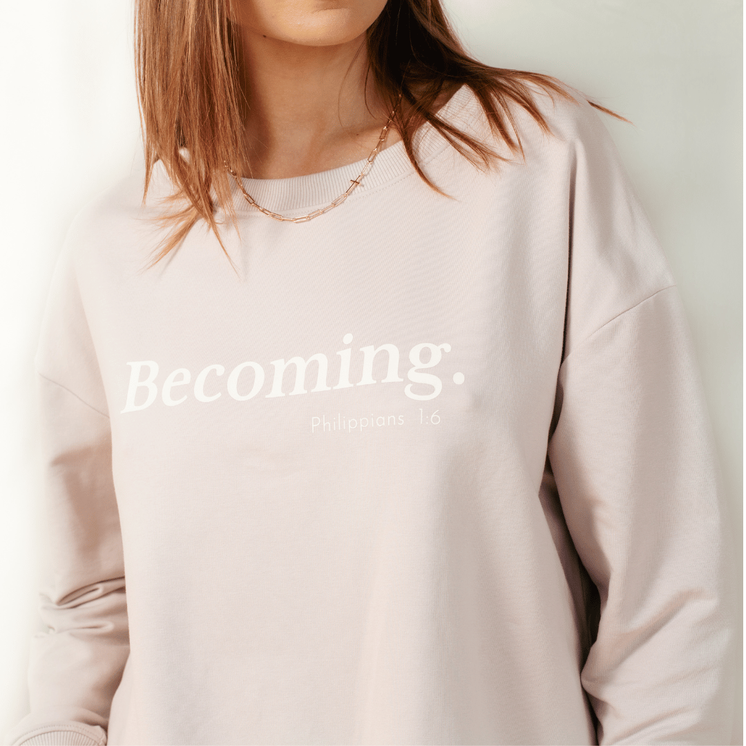 Christian woman wearing the shore beige "Becoming. Philippians 1:6" Peruvian cotton crew sweatshirt from the Be The Light Collection by Rizen Jewelry.